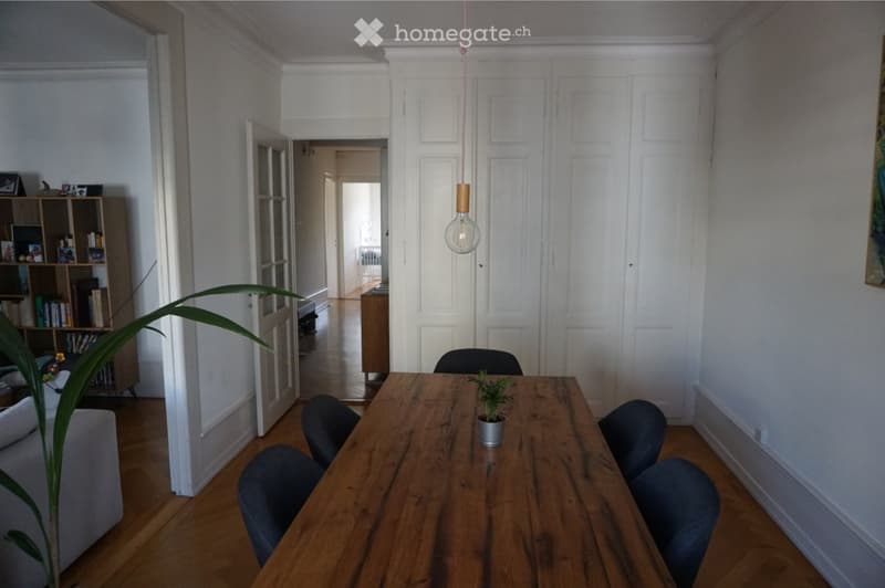 A well-lit dining area with a wooden table, six chairs, a sofa, a bookshelf, and a potted plant. The room features light-colored walls, wooden flooring, and a hanging pendant light.