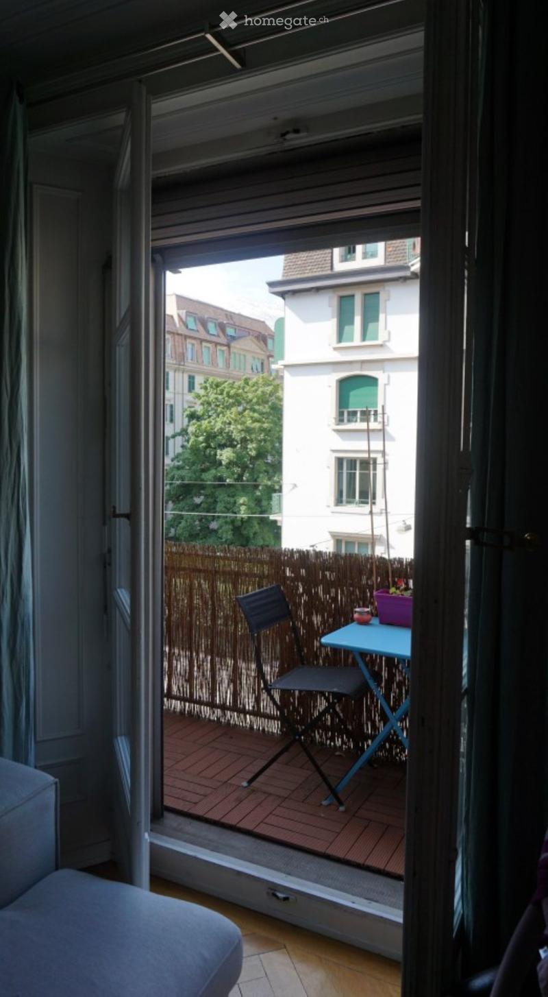 Balcony with wooden decking, a small table, a folding chair, and a flower pot, overlooking a cityscape with buildings and greenery.