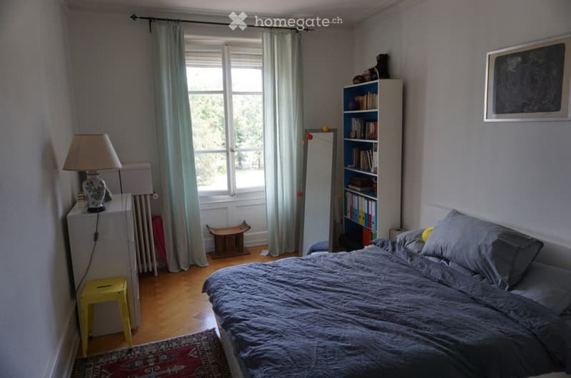 Cozy bedroom with a large bed, nightstand with lamp, bookshelf, mirror, and a small yellow stool near the window.