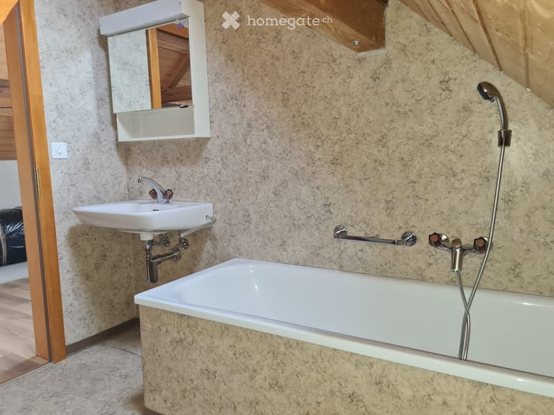 Modern bathroom with a bathtub, sink, mirror, and faucet. The walls are covered in beige stone tiles, and the floor is tiled. A wooden door is visible on the left side.