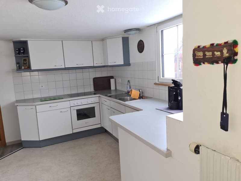 A modern kitchen with white cabinets, a stainless steel sink, an oven, and a coffee maker on the counter. The space is well-lit with natural light from a window and features tiled walls and a light-colored floor.