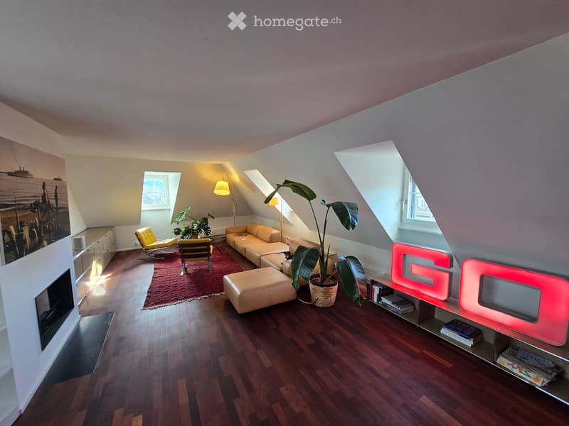 Modern living room with wooden flooring, slanted ceiling, large windows, and skylights. Features a cream-colored sofa, ottoman, yellow chair, floor lamp, potted plants, and a red neon sign on a shelf.