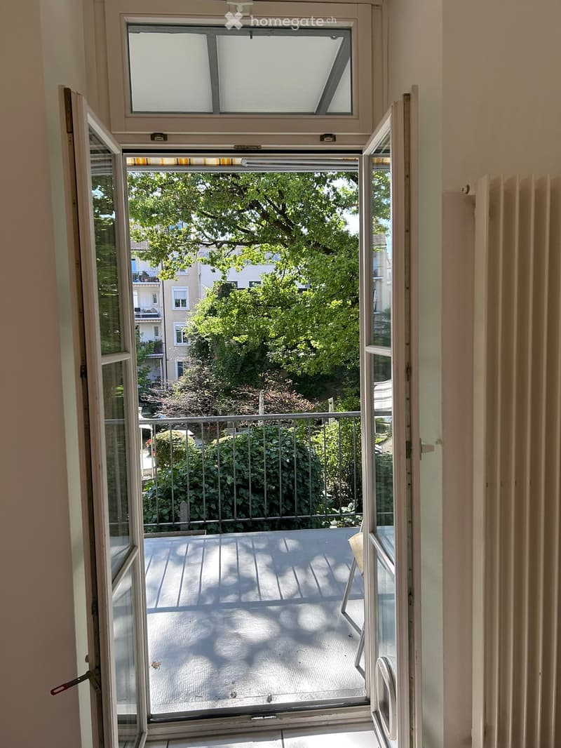 Open sliding glass door leading to a small balcony with wooden decking, metal railing, and greenery visible outside.