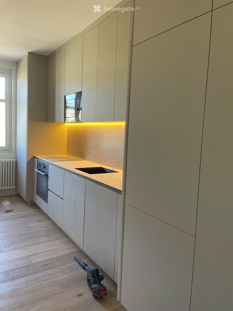 Empty kitchen with light-colored cabinets, a countertop, sink, and built-in oven. Wooden flooring and a window on the left side.