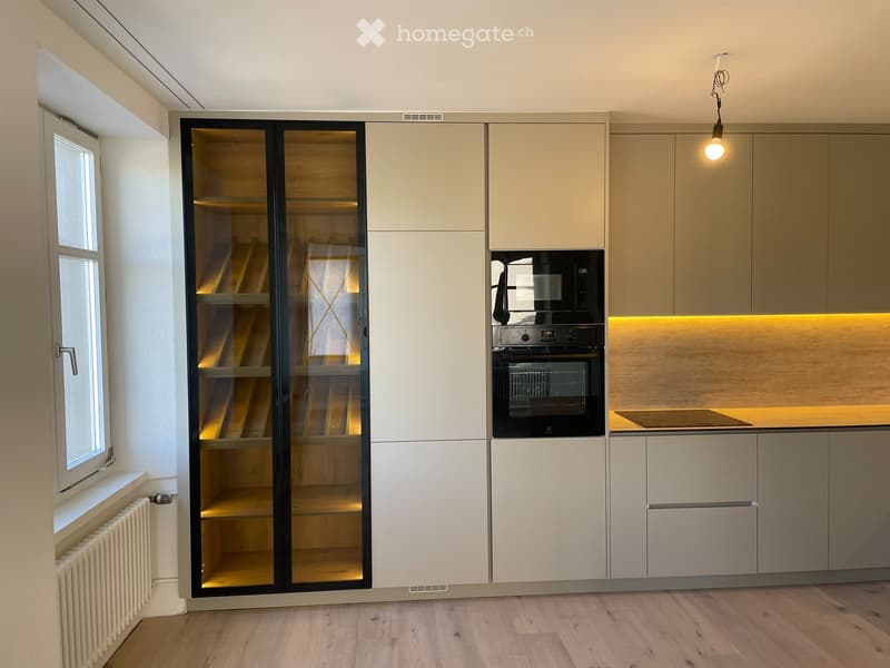 Modern kitchen with light-colored cabinets, glass-fronted storage, built-in oven, and warm lighting.