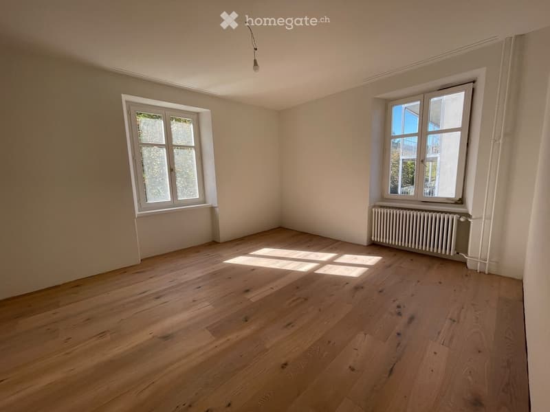 Empty room with light wooden flooring, white walls, and two windows allowing natural light to enter. A radiator is visible under one window.