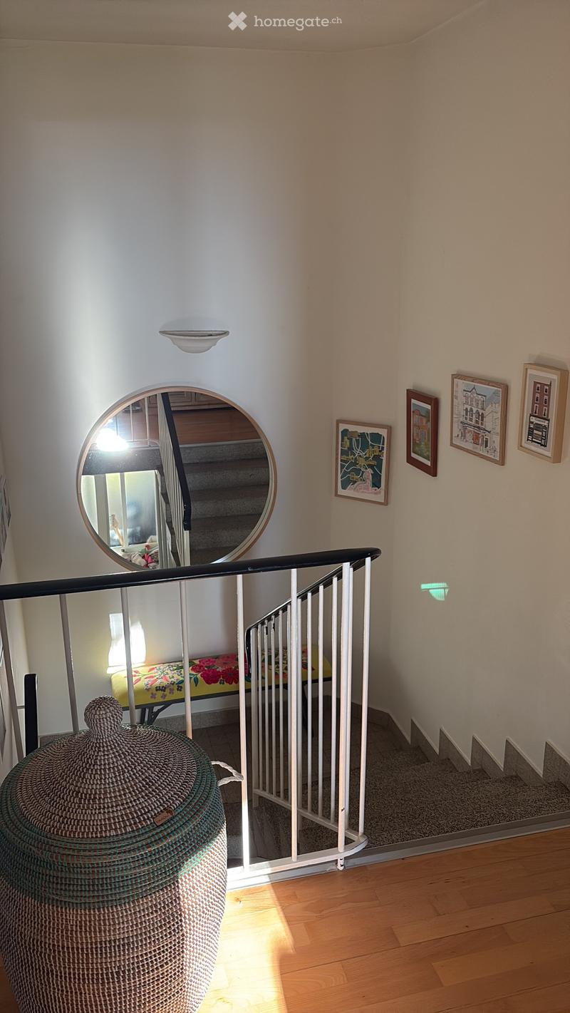 Staircase with wooden steps, white railing, and a round mirror on the wall. The hallway features framed artwork and a wicker basket on the floor.