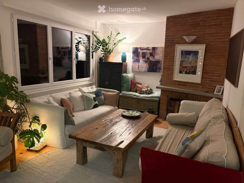 Living room with a white sectional sofa, wooden coffee table, armchair, and decorative pillows. Features large windows, a brick accent wall, and artwork.