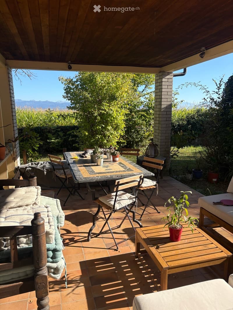 A covered outdoor terrace with a tiled floor, wooden furniture including a sofa, coffee table, dining table, and chairs, surrounded by greenery and offering a scenic mountain view.