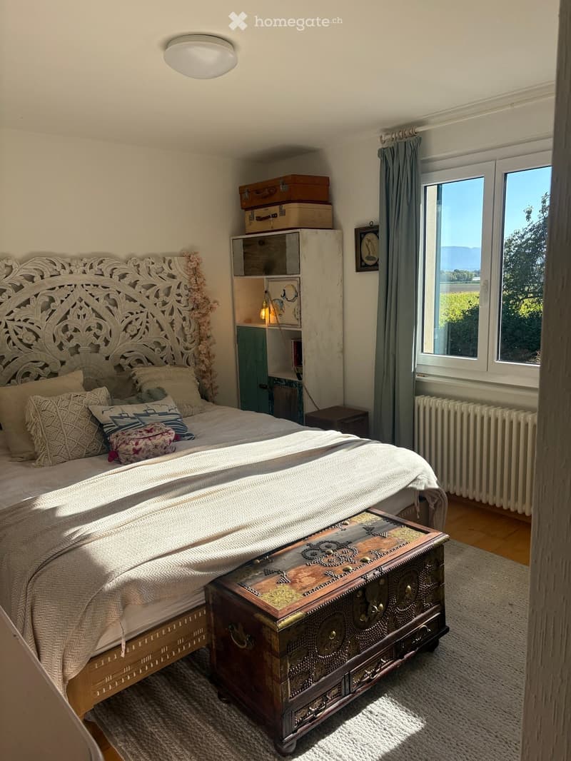 Cozy bedroom with a large, intricately carved wooden bed, patterned bedding, a vintage trunk as a nightstand, a wooden wardrobe, and a scenic view through large windows.