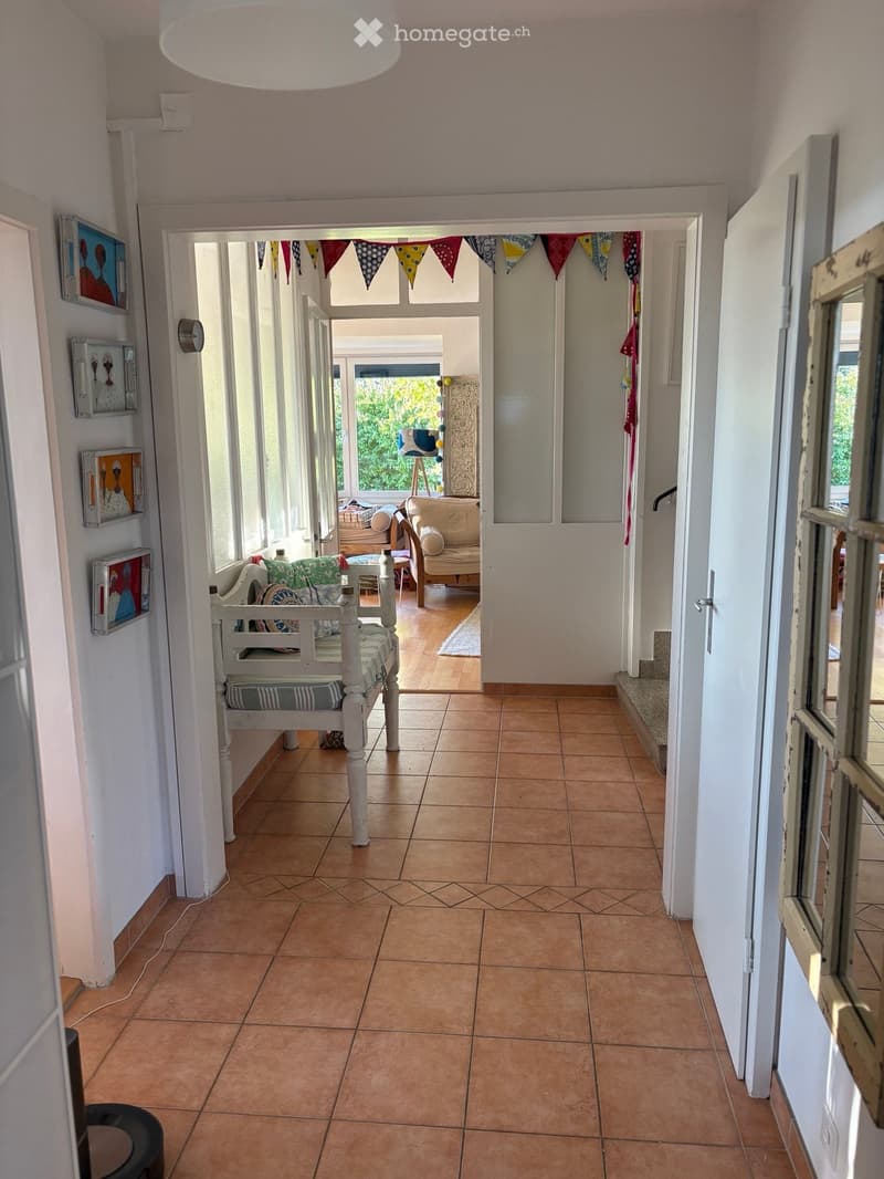 Bright hallway with tiled floor, white walls, and a bench. The hallway leads to a living area with a sofa, armchair, and decorative items.