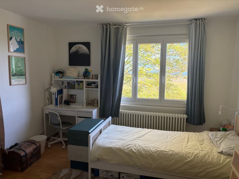 Cozy bedroom with a single bed, desk, chair, and shelves. Large windows with grey curtains let in natural light. The room features a radiator, framed pictures on the wall, and a small suitcase on the floor.