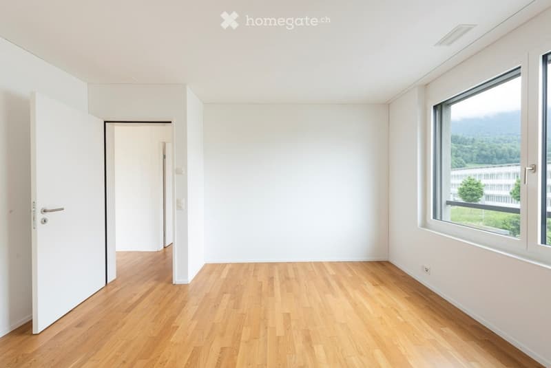 Empty room with light wooden flooring, white walls, and large windows offering a scenic view. The room features a door leading to another space.