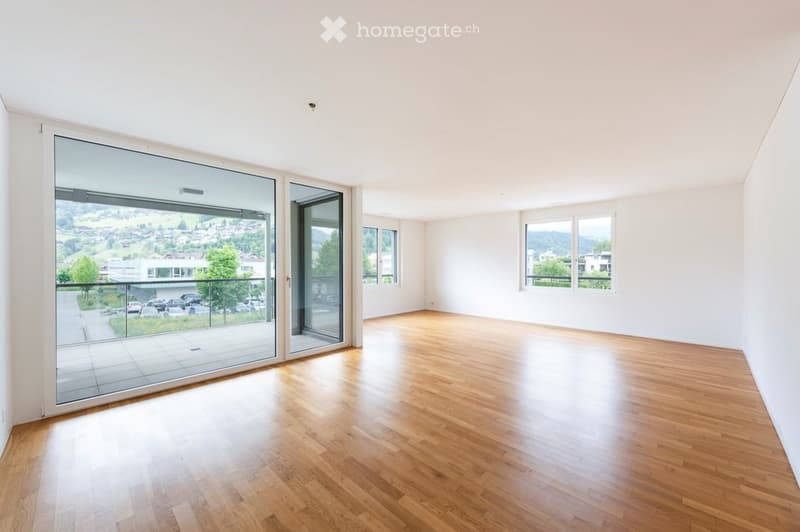 Empty room with wooden flooring, white walls, large glass sliding doors leading to a balcony, and windows offering a scenic view of the surrounding area.