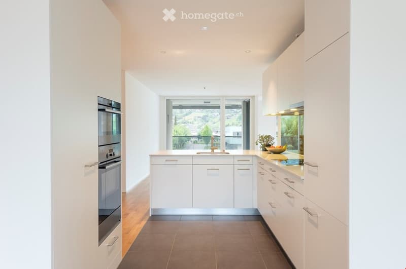 Modern kitchen with white cabinets, stainless steel oven, central island, and a view through large windows.
