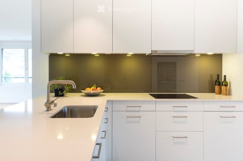 Modern kitchen with white cabinets, stainless steel sink, and countertop. Features a bowl of fruit, a potted plant, and three bottles on the counter.