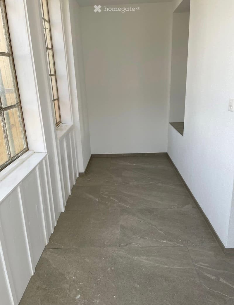 Empty hallway with white walls, dark tiled floor, and a window on the left side.
