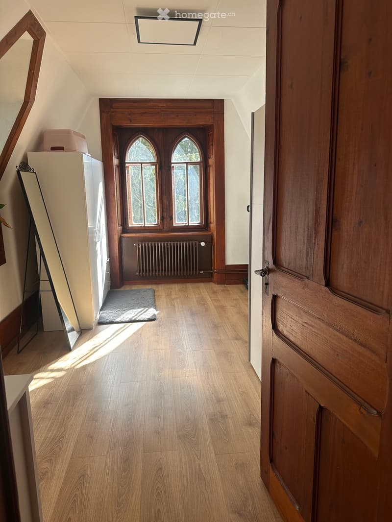 A narrow hallway with wooden flooring, arched windows at the end, and a radiator below them. A small rug is placed on the floor, and a mirror leans against the wall on the left side.