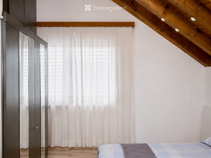 Cozy attic bedroom with wooden slanted ceilings, white walls, and large window with sheer curtains. Features a neatly made bed with a blue and grey checkered duvet and matching pillows, and a modern grey wardrobe with mirrored doors.