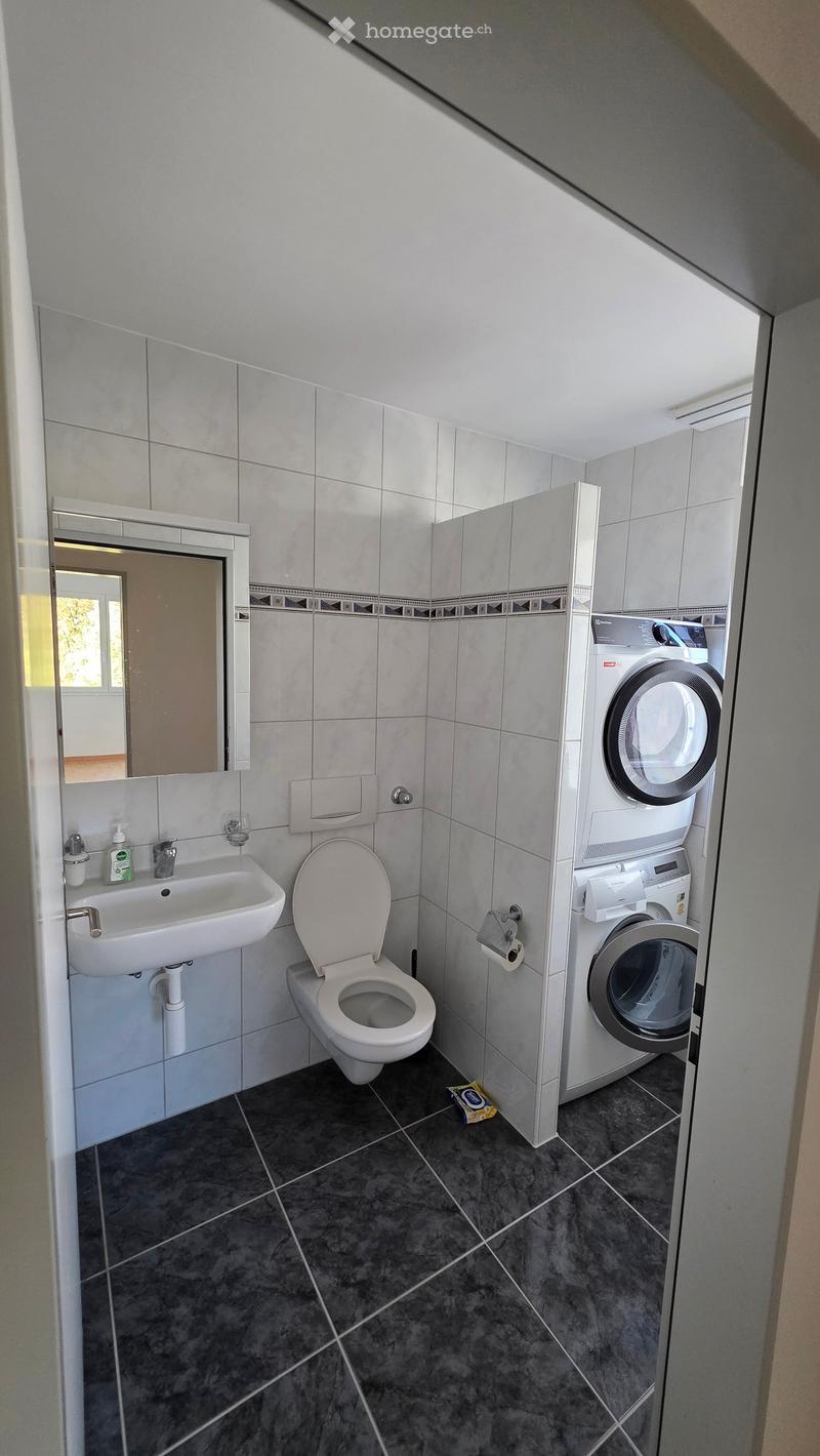 Modern bathroom with white tiles, a sink, toilet, mirror, and a stacked washing machine and dryer unit. The floor is dark grey tiled.