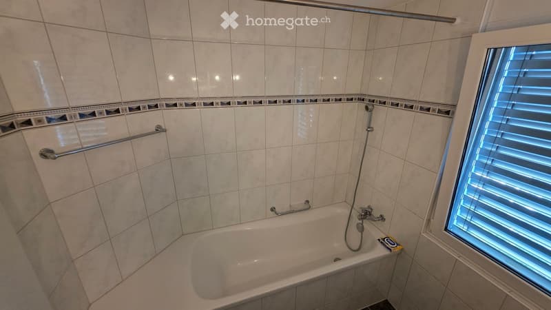 Bathroom with a bathtub, tiled walls, a window with blinds, and a handrail for support.