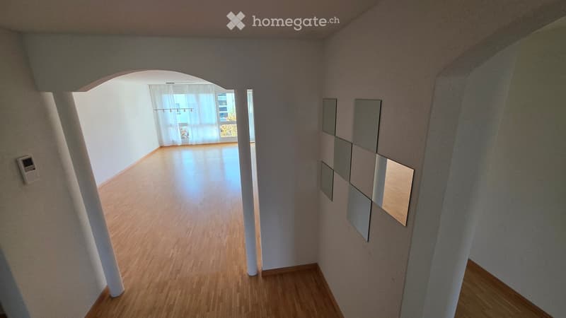 Empty hallway with wooden flooring, white walls, and a decorative wall panel on the right side. An open doorway leads to a bright room with large windows and sheer curtains.