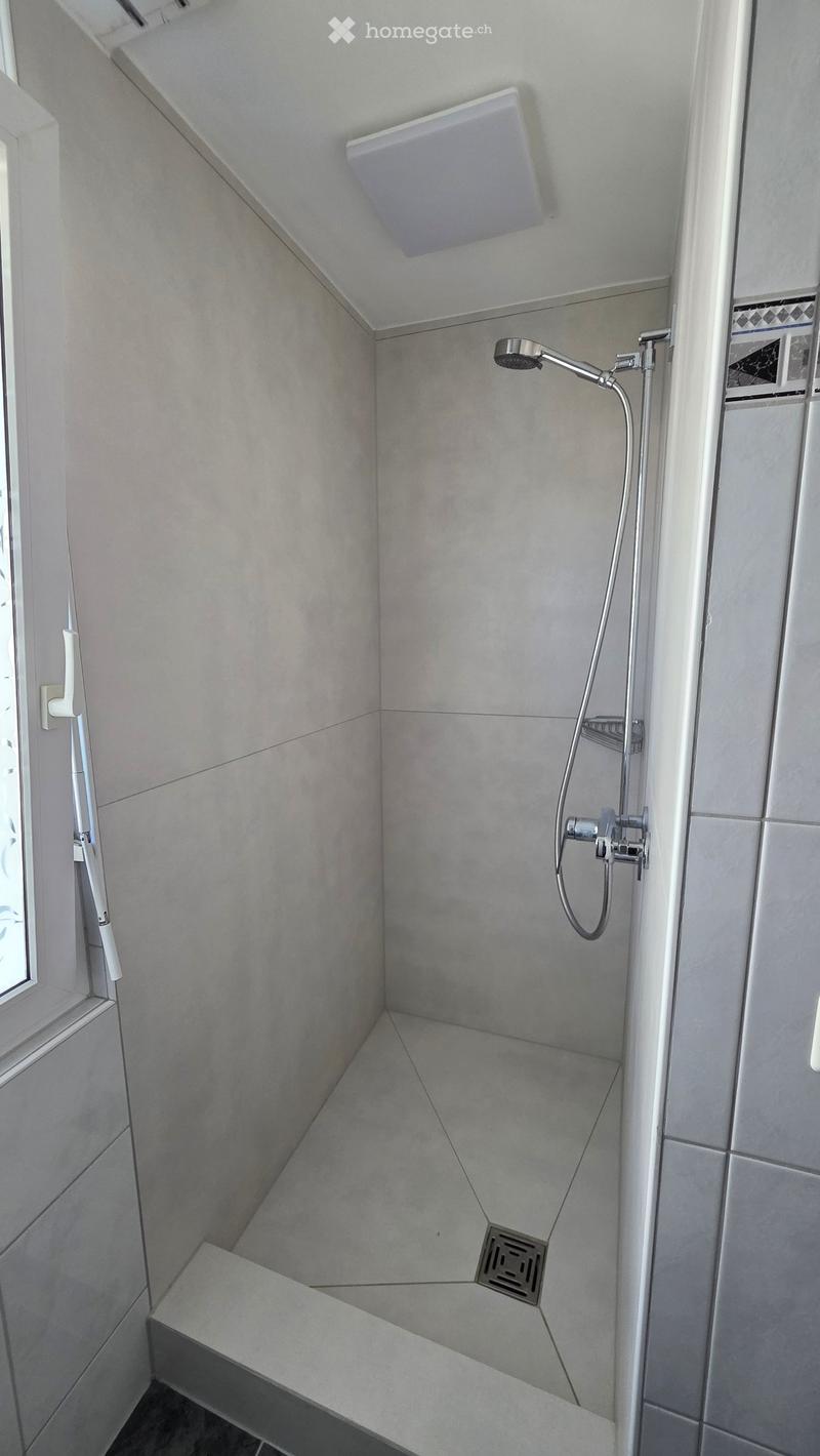 Modern shower area with grey tiled walls and floor, a handheld showerhead, and a built-in seat.