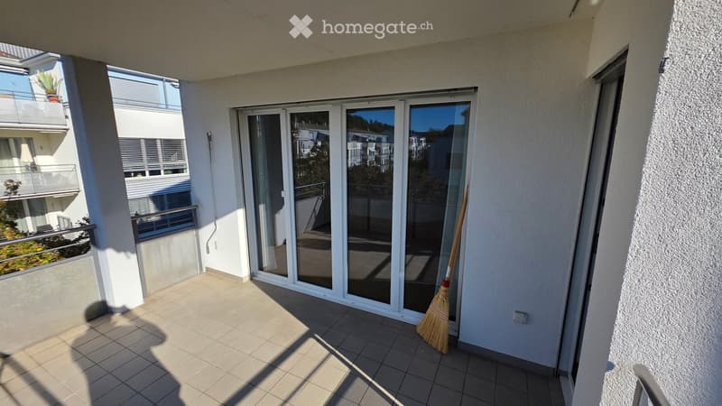 Balcony with tiled floor, large sliding glass doors, and a broom leaning against the wall. The balcony overlooks neighboring buildings and greenery.
