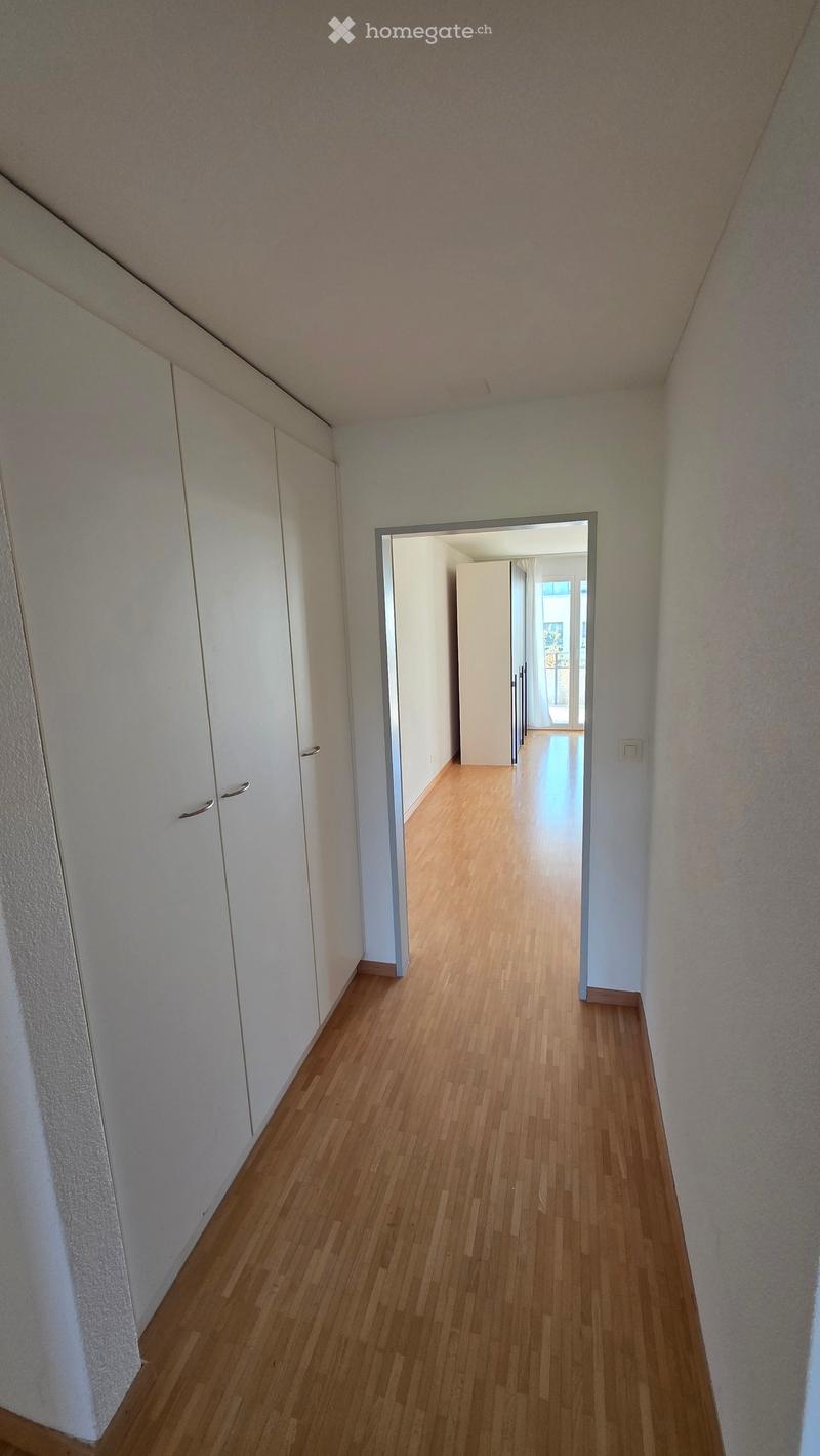 A narrow hallway with wooden flooring, white walls, and built-in white cabinets on one side. The hallway leads to an open doorway revealing another room with light-colored walls and wooden flooring.