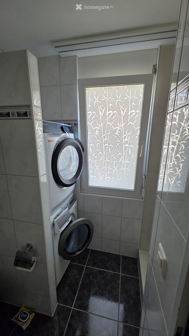 Bathroom with tiled walls and floor, featuring a washing machine and dishwasher unit, a small window with decorative glass, and a toilet paper holder.
