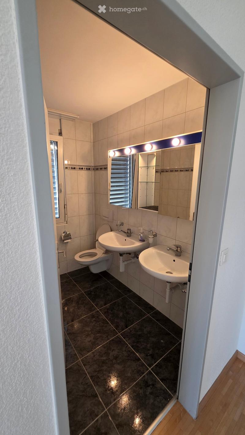 Modern bathroom with dark tiled floor, white tiled walls, a toilet, two sinks, a large mirror with built-in lighting, and a window with blinds.
