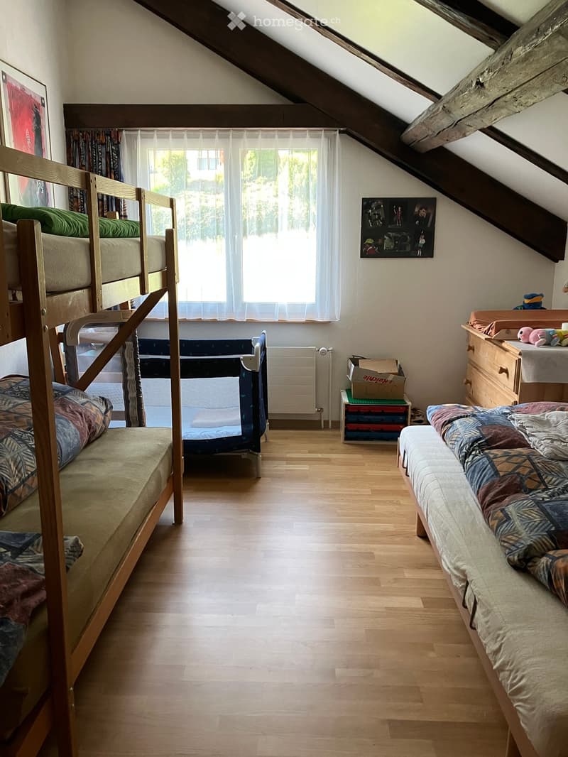 Cozy attic bedroom with wooden floors, bunk bed, single bed, nightstand, and toys. Large window with curtains and a radiator below.