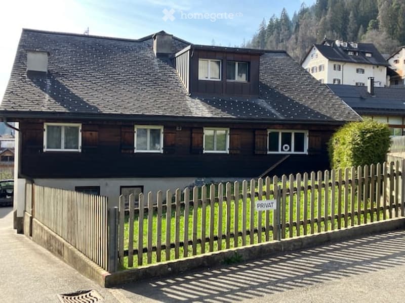 Two-story house with dark wooden siding and white-framed windows, featuring a pitched roof and a wooden fence in front.