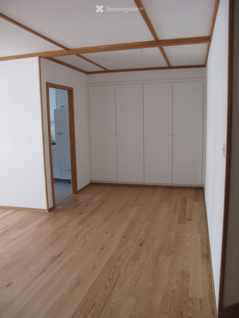 Empty room with light wooden flooring, white walls, and built-in white closet units. The ceiling features exposed wooden beams.
