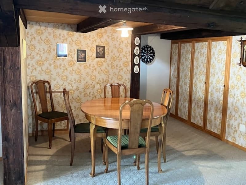 A cozy dining area with a round wooden table surrounded by six wooden chairs with green cushions. The walls are decorated with framed pictures and wall plates, and there is a dartboard mounted on the wall.