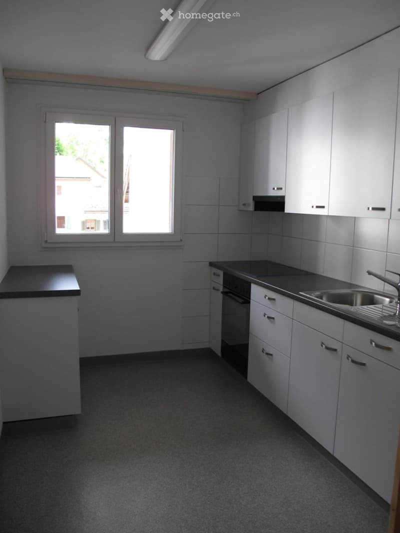 Modern kitchen with white cabinets, black countertops, a sink, and an oven. The room has large windows and a grey floor.