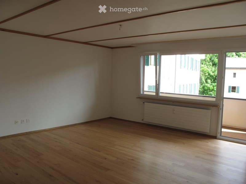Empty room with wooden flooring, white walls, and large windows with a view of greenery outside.