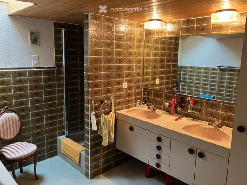 Bathroom with double sink, mirror, tiled walls, and a small chair near the shower area.