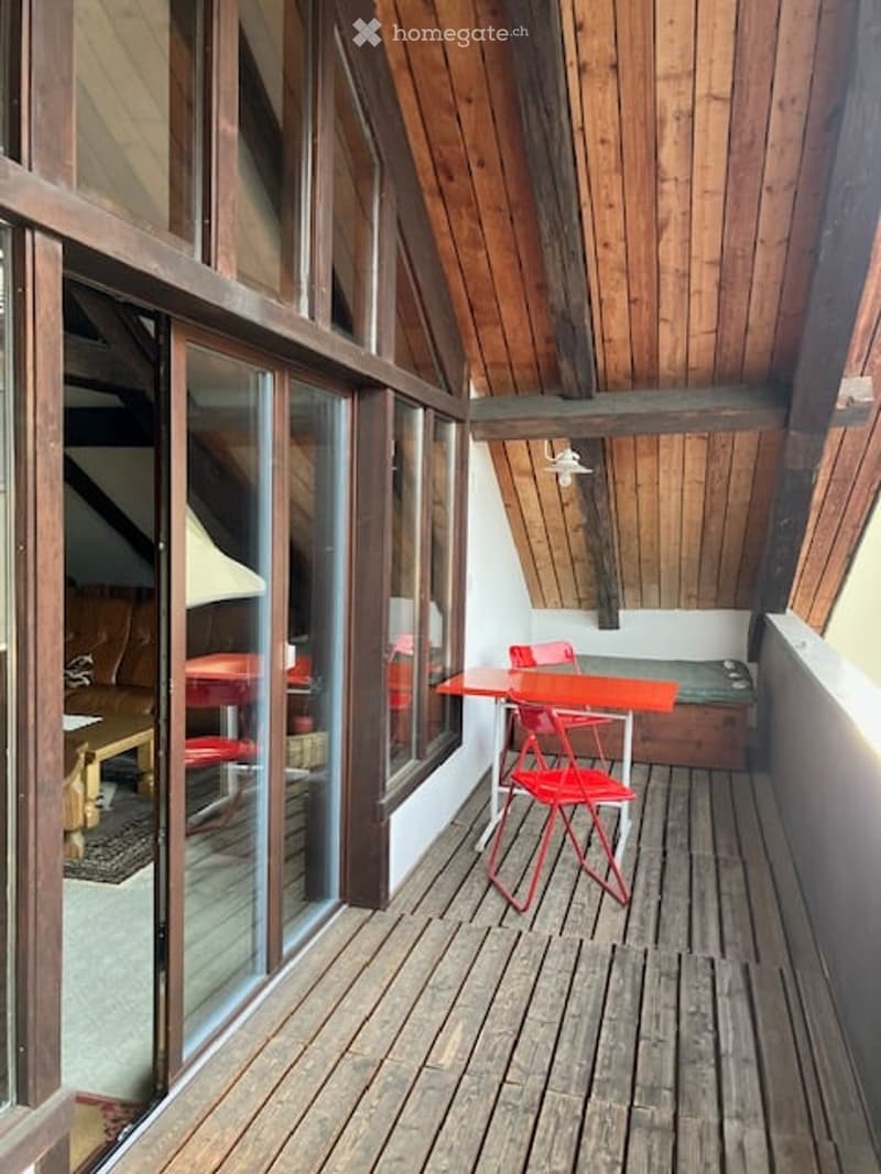 Wooden balcony with large windows, red chairs, and a small table.