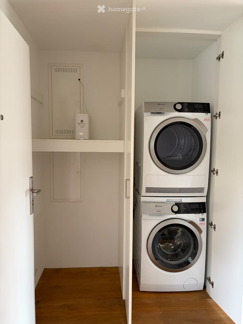Compact bathroom with a stacked washing machine and dryer unit, wooden flooring, and built-in storage cabinets.