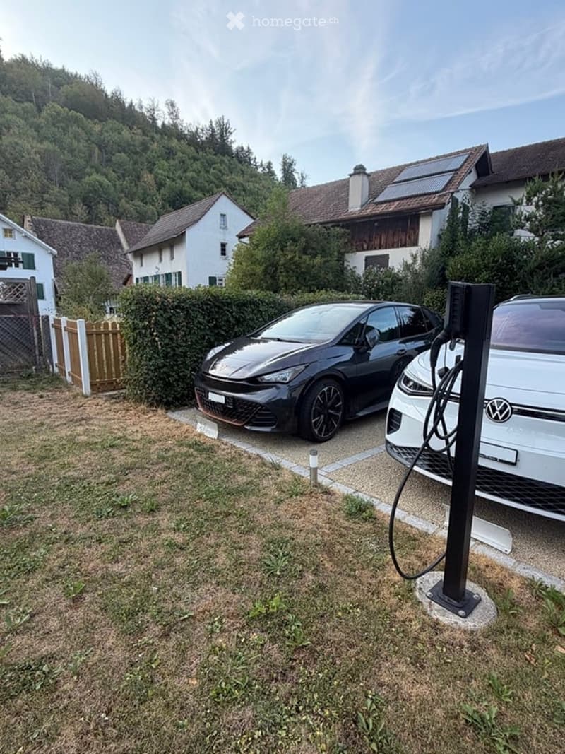 Residential area with two parked cars, one connected to an electric charging station, surrounded by houses and greenery.