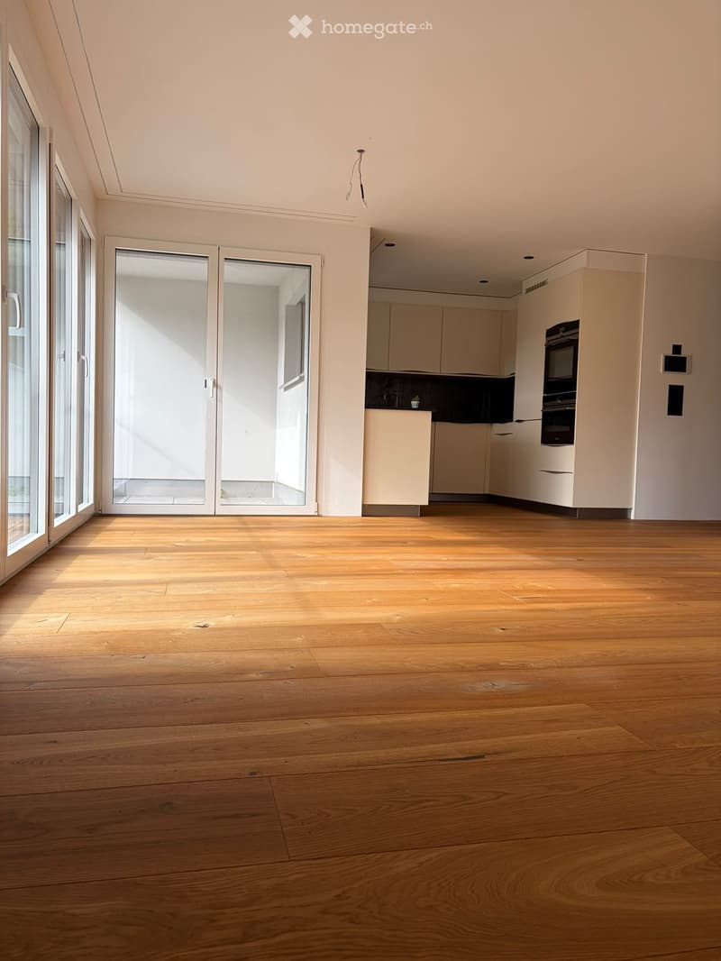 Empty room with wooden flooring, large windows, and a small kitchen area with cabinets and appliances visible in the background.
