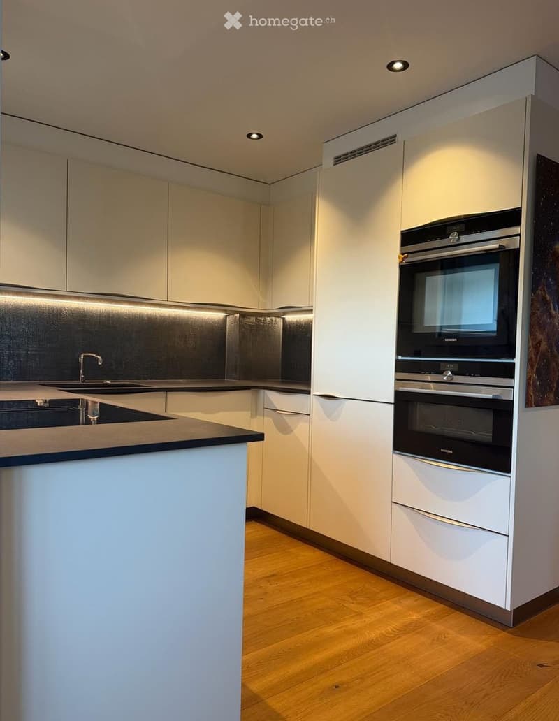 Modern kitchen with white cabinets, stainless steel appliances, and a dark countertop. Features include a built-in oven, a sink with a modern faucet, and sleek lighting.