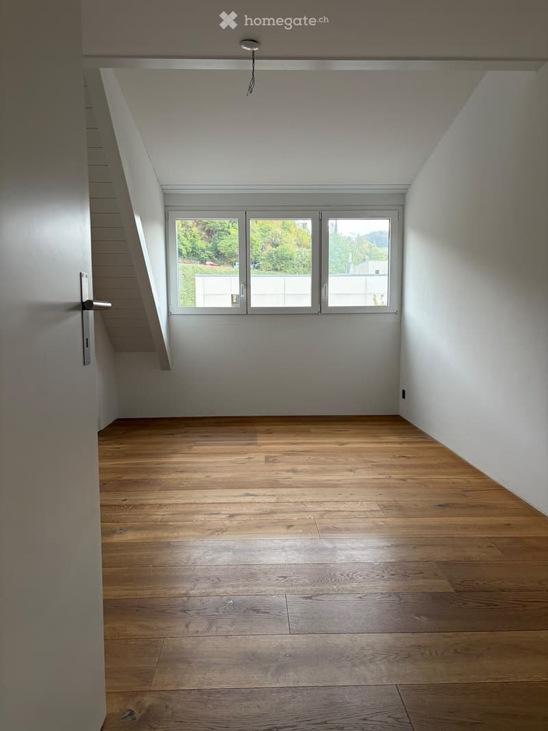 Empty room with wooden flooring, white walls, and large windows offering a view of greenery outside.