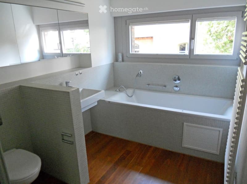 Modern bathroom with a bathtub, sink, toilet, and large windows. Features grey tiled walls, wooden flooring, and a radiator.