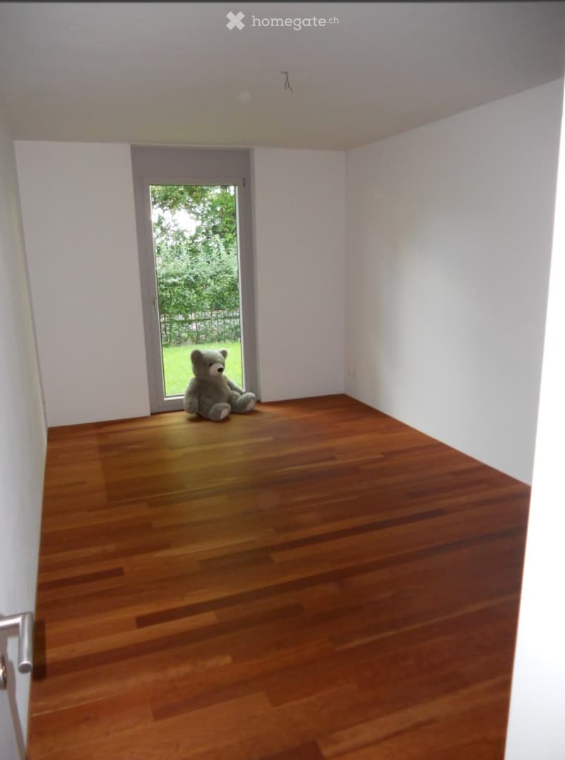 Empty room with wooden flooring, white walls, and a large window with a view of greenery outside.