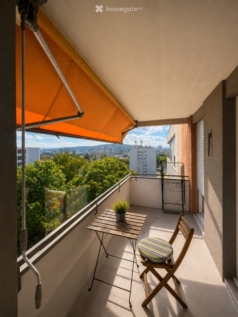 Balcony with a wooden table, chair, and small potted plant, featuring a retractable awning and a view of greenery and cityscape.