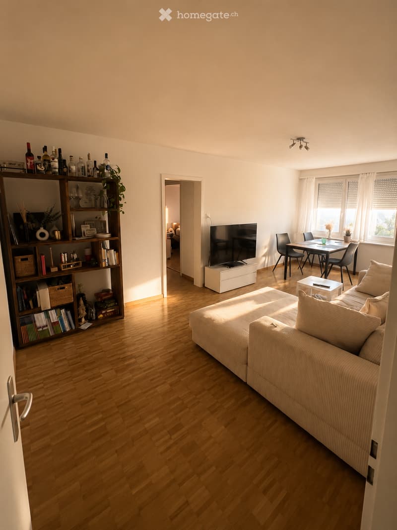 Bright living room with wooden flooring, a beige sofa, coffee table, bookshelf, dining table with chairs, and a television on a stand.