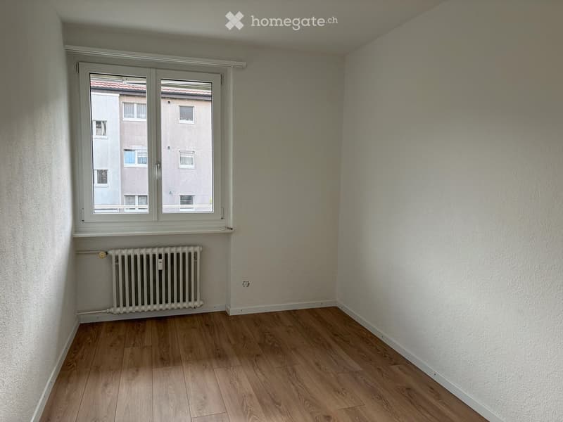 Empty room with wooden flooring, white walls, a window with a view of neighboring buildings, and a radiator below the window.
