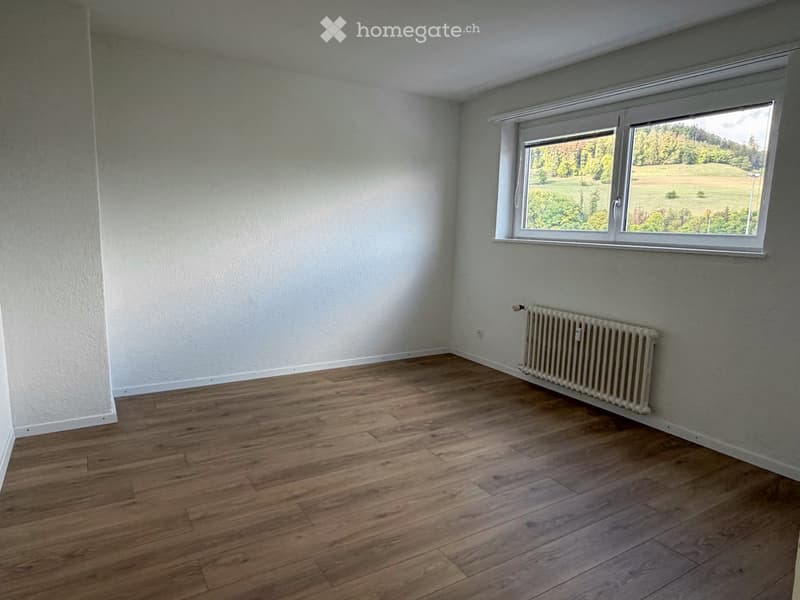 Empty room with wooden flooring, white walls, a single window with a view of greenery, and a radiator below the window.